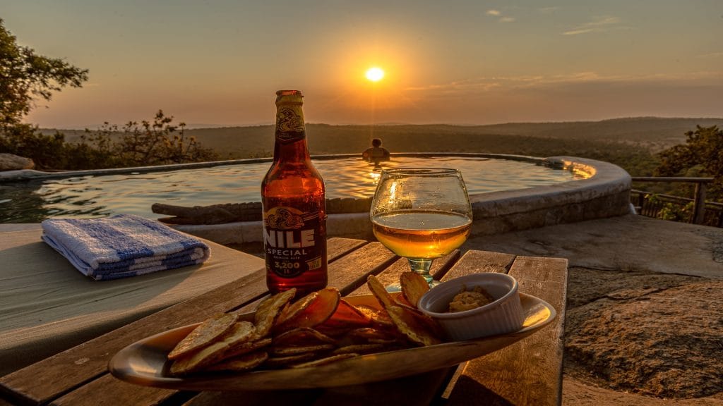 Bottle of Nile Special beer with a glass and snacks on a table beside a swimming pool at a Uganda safari lodge. File Name